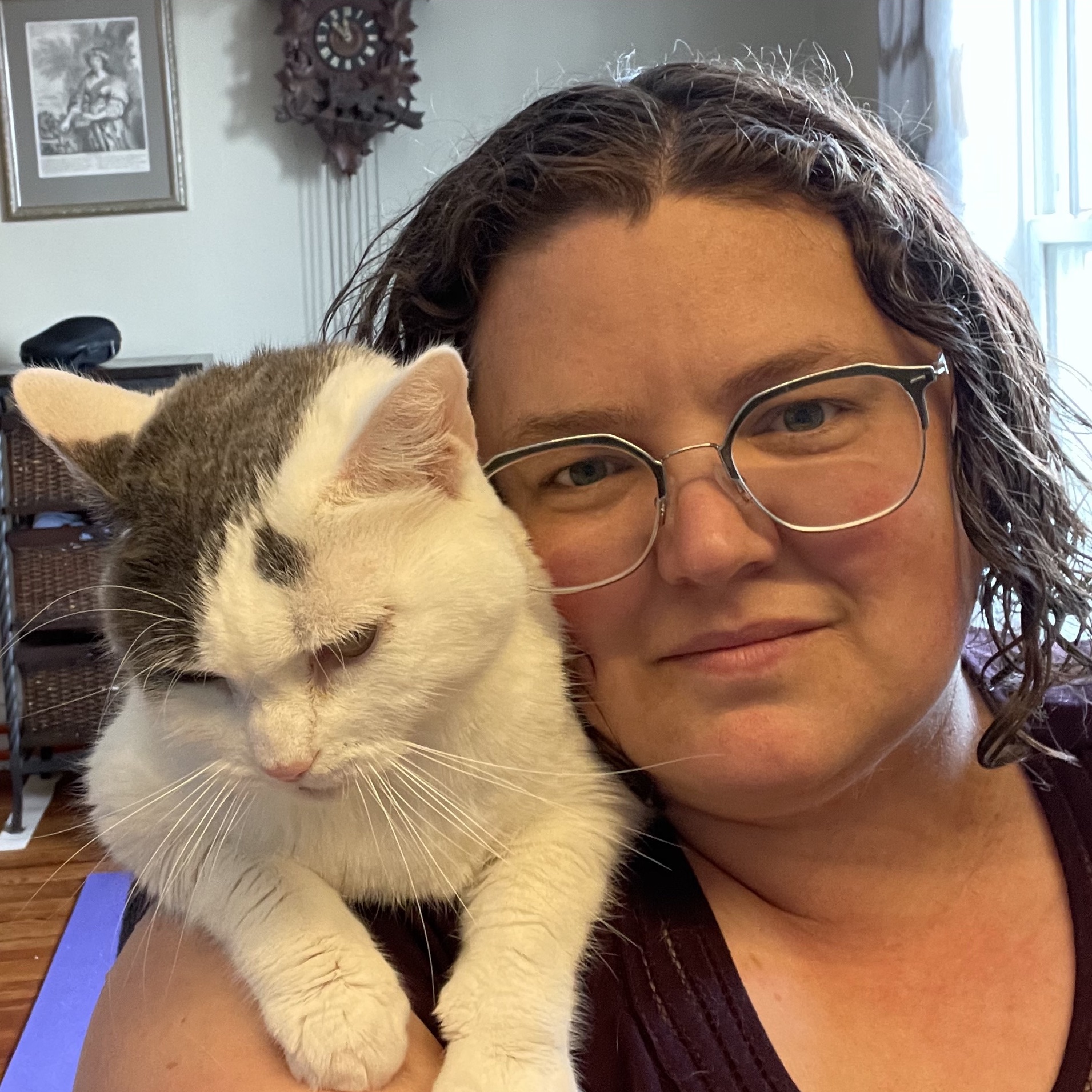 A smiling white woman with glasses, rosy cheeks, and curly brown hair. Her pretty white-and-grey cat is on her shoulder.