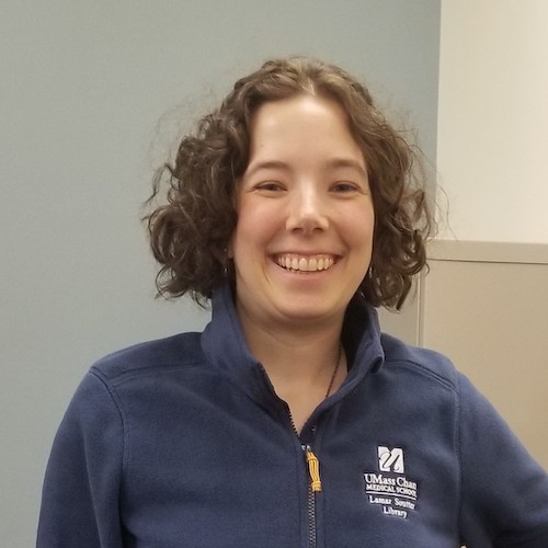 Tess Grynoch, a white woman with short brown curly hair, smiling and proudly wearing her UMass Chan Medical School, Lamar Soutter Library sweater.