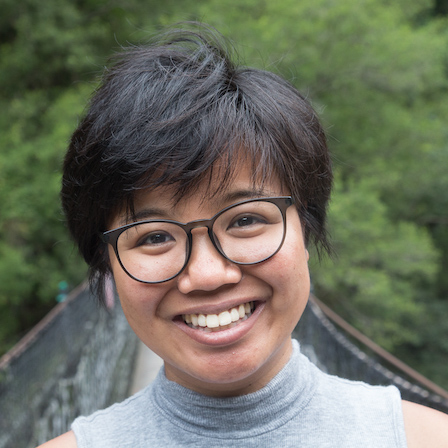 A medium-skinned Asian woman with a pixie cut wearing a gray shirt and round eyeglasses with black frames. Trees and other greenery are behind her.