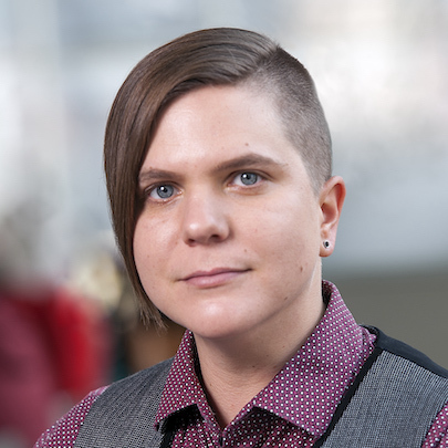 An androgynous white woman with a subtle smile, asymmetrical light brown hair shaved on the left side, wearing a dark red shirt with a white pattern and grey/black vest. The background blurred.