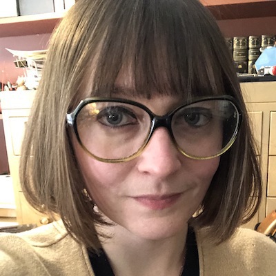 White female with short light brown hair wearing green glasses, a gold necklace, and a beige cardigan smiles. A bookcase and old card catalog are behind her.